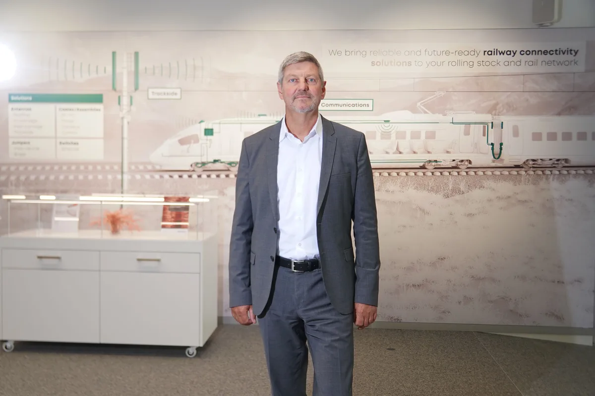 Porträt von Urs Ryffel. Der Mittfünfziger trägt einen grauen Anzug und ein weisses Hemd und steht vor einer Wand, auf der eine Zugkomposition abgebildet ist.